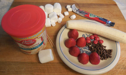 Frosting, marshmallows, strawberries, chocolate chips, m&ms, a banana and fruit roll up on a cutting board.
