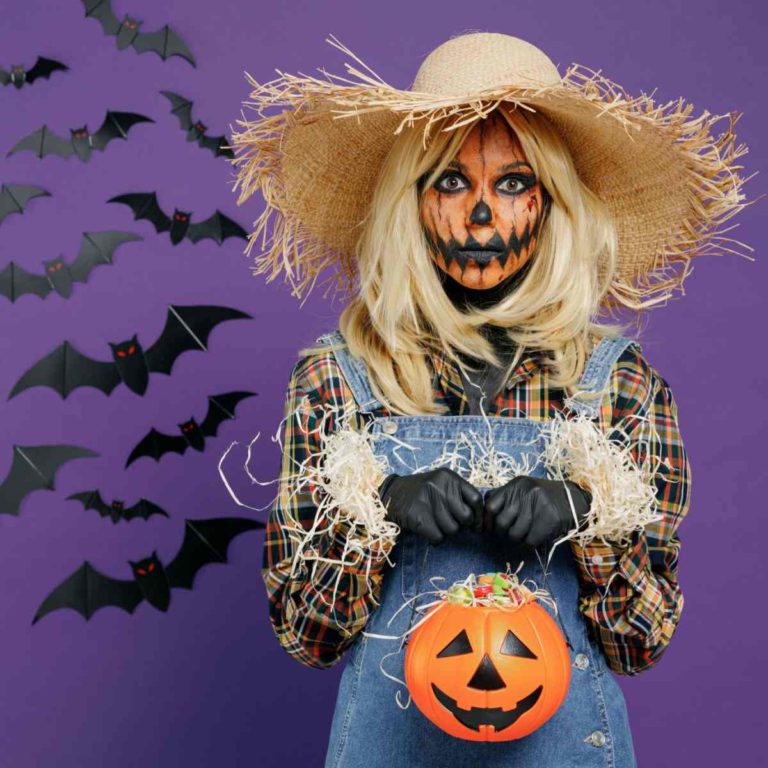 A woman wearing a DIY scarecrow costume for Halloween, holding a plastic jack-o-lantern filled with candy, standing against a purple wall with paper bats on it.