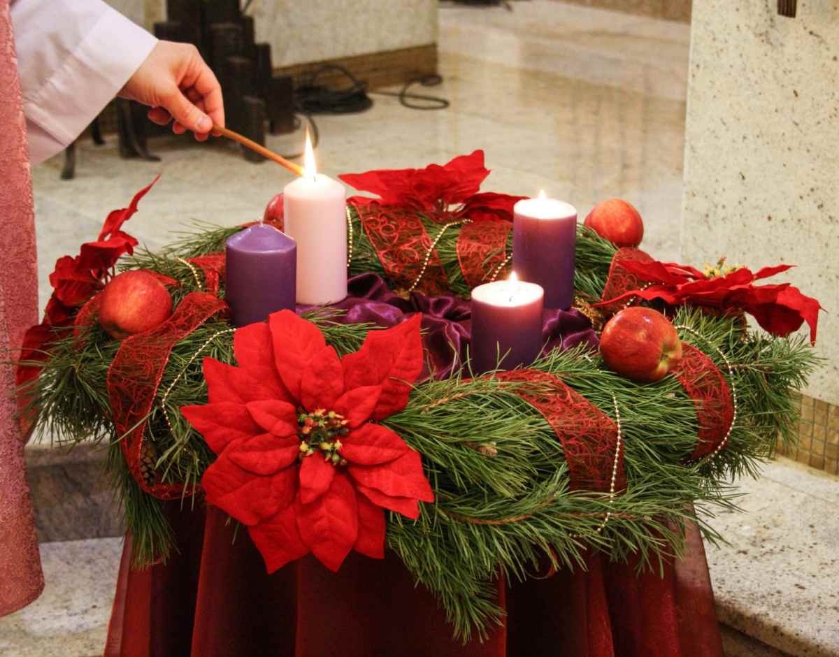 A type of Christmas wreath called an Advent Wreath, which has red ribbon wrapped around it, with a poinsettia and red ornaments on it, and four candles in the center - three of which are purple, and the fourth is light pink, and there is a woman's hand lighting it with a long match.