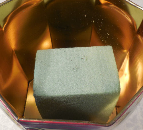 Floral foam sitting inside a decorative tin showing the first step of a craft project.