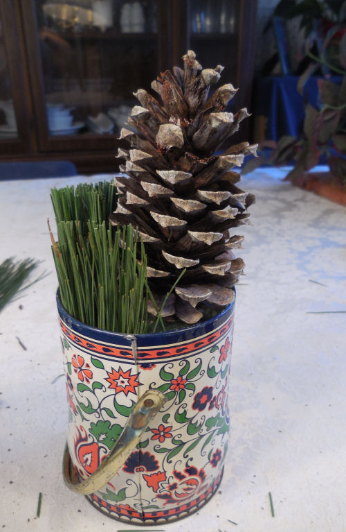 A decorative tin with a pine cone and pine needles on a countertop.
