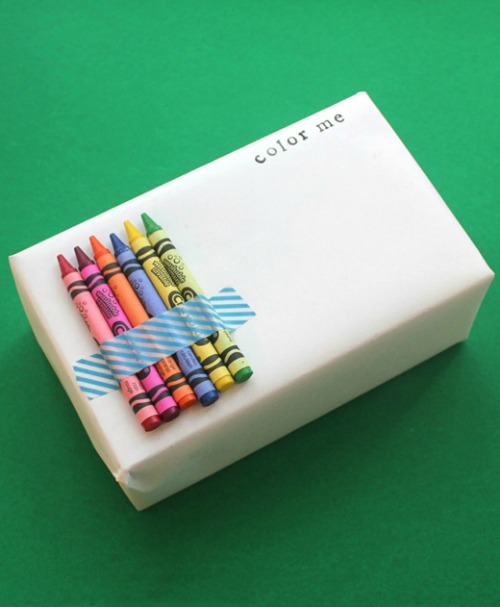 A present wrapped in white paper with the words "color me" written on it and six Crayons on it. 