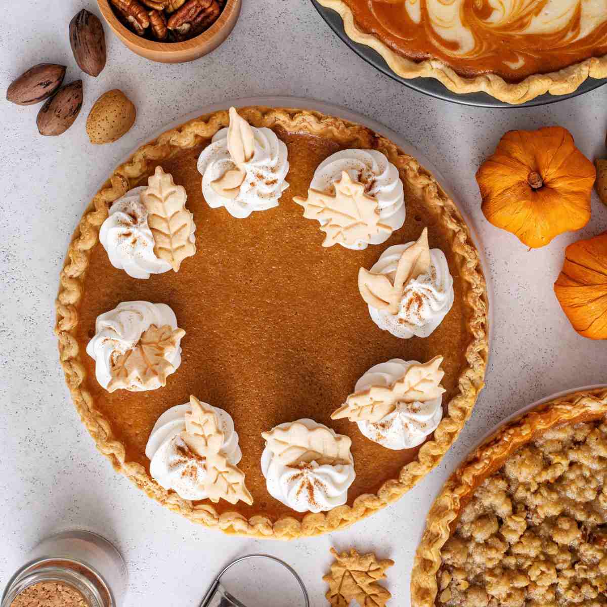 A homemade pumpkin pie with dollops of whipped cream and leaf cookies on it, with two mini pumpkins to the right of it, and around the border, two pumpkins and some spices are also seen.