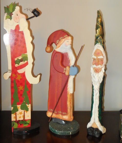 Three tall Santa figures, one green, one red, and one with holly in his hair smoking a pipe..