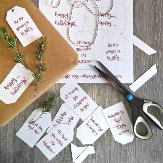 Holiday gift tags with messages on a table next to scissors, rosemary and a wrapped present.