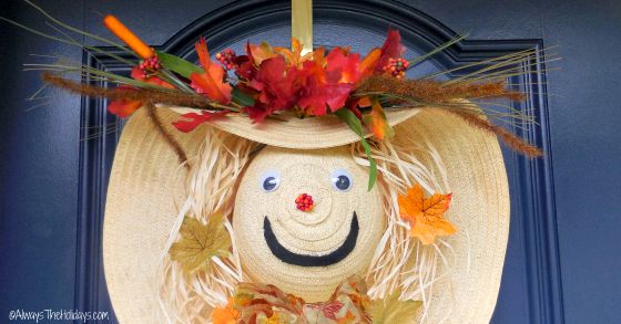 Straw hat scarecrow wreath A close up image of a straw hat scarecrow wreath with raffia hair, and fall leaves hanging on a blue door with a gold door hanger.