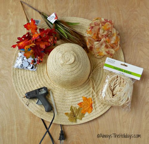 supplies for the scarecrow autumn wreath Supplies for scarecrow wreath door decoration including fall floral picks, orange wire ribbon, natural raffia, google eyes and a hot glue gun sitting on top of a floppy wide brimmed straw hat that's on a table.