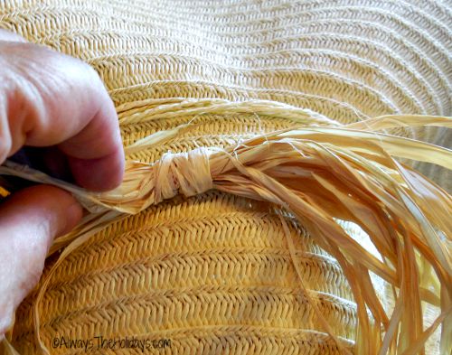 A piece of natural raffia attached wit hot glue to a scarecrow straw hat, with a woman's hand holding one side of the raffia. 