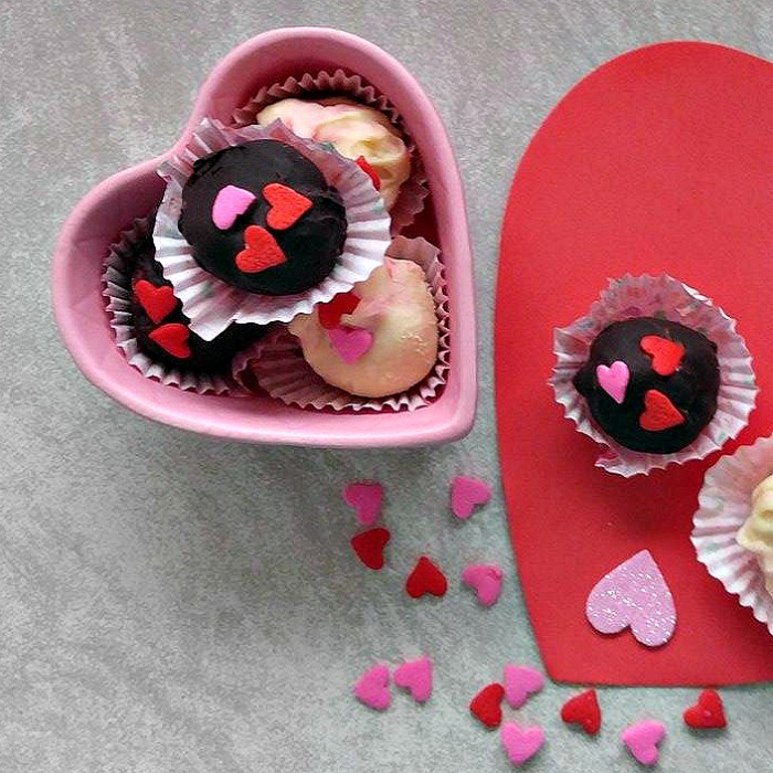 Raspberry cheesecake truffles in a heart shaped bowl next to candy heart sprinkles on top of a large red paper heart with two additional truffles on top.