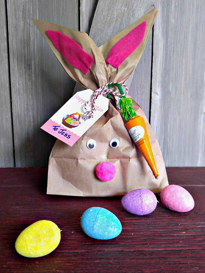 An Easter Bunny gift bag that is in the shape of a bunny, with an Easter gift tag and candy tied to it, and Easter eggs in the foreground of the photo.
