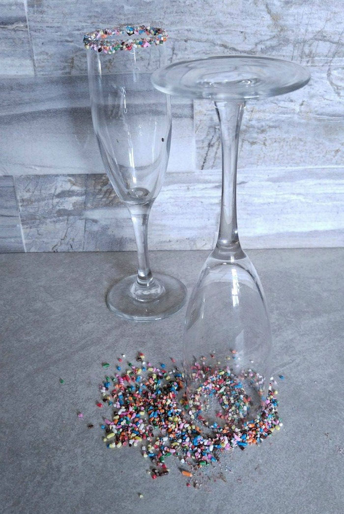 Glass rims dipped in sprinkles. Two champagne flutes, one is right side up and one is upside down to dip the rim of the glasses in sprinkles.