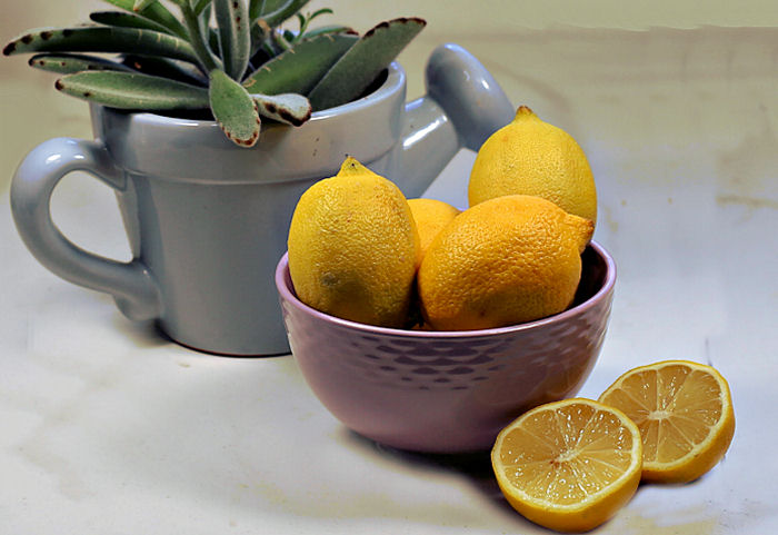 A bowl of lemons. A pink bowl of lemons next to a plant in a grey watering can shaped pot on a counter.