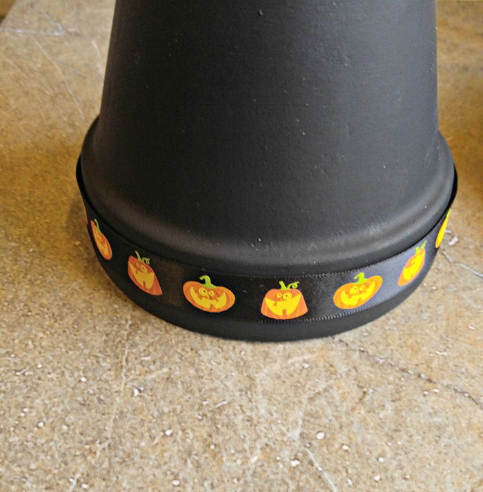 Attach the ribbon to the pot rim. Black Halloween ribbon with pumpkins on it glued to the rim of a painted black clay pot.