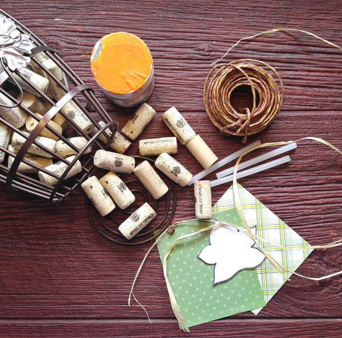Materials for DIY wine cork pumpkin craft including wine corks, heavy card stock paper, glue sticks, raffia, wired jute, and orange acrylic craft paint.