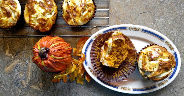 Pumpkin cheesecake muffins on a plate A small oval plate with two pumpkin cheesecake muffins on it, next to a faux pumpkin and a wire rack with more pumpkin cheesecake swirl muffins.