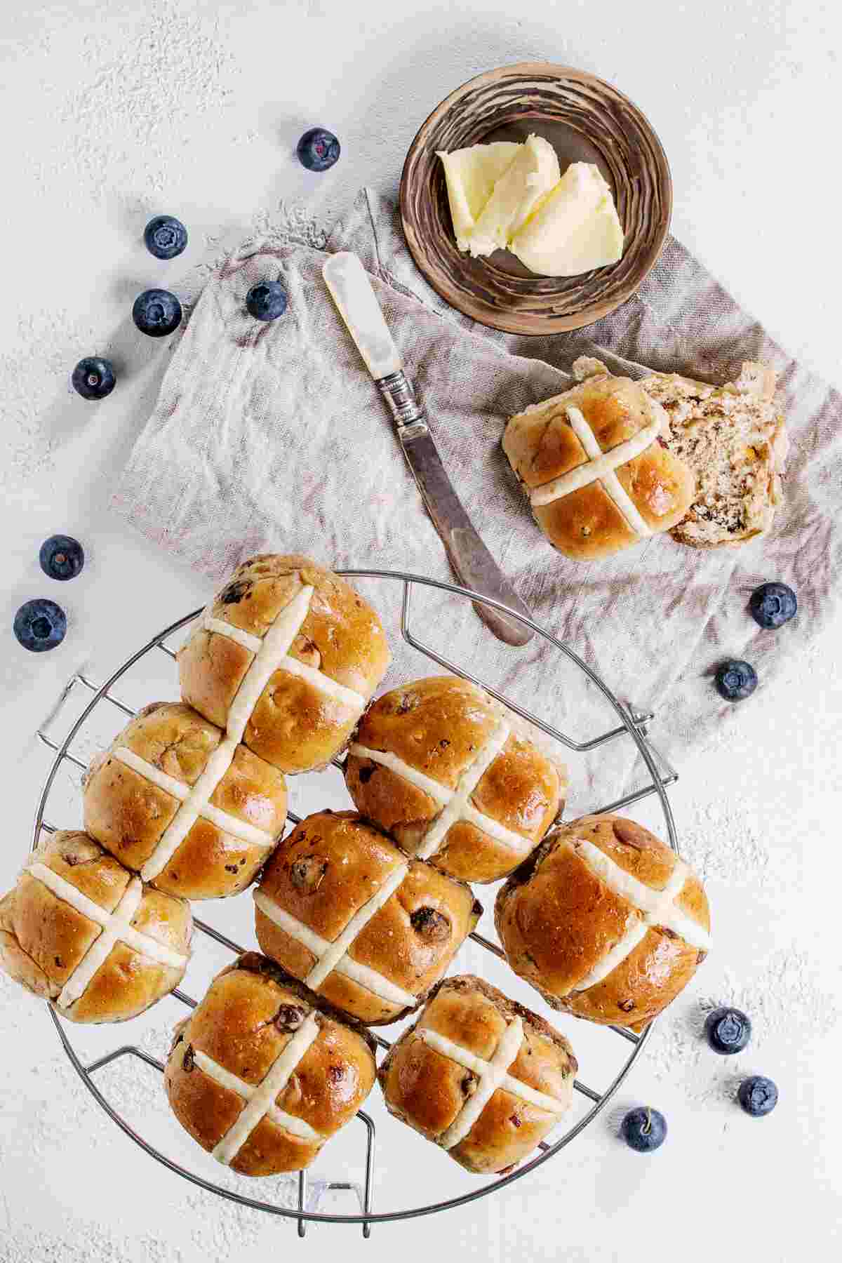 A baking rack of homemade hot cross buns, a traditional Easter bread, above a neutral linen napkin, with a pat of butter in a ramekin and blueberries scattered around it.