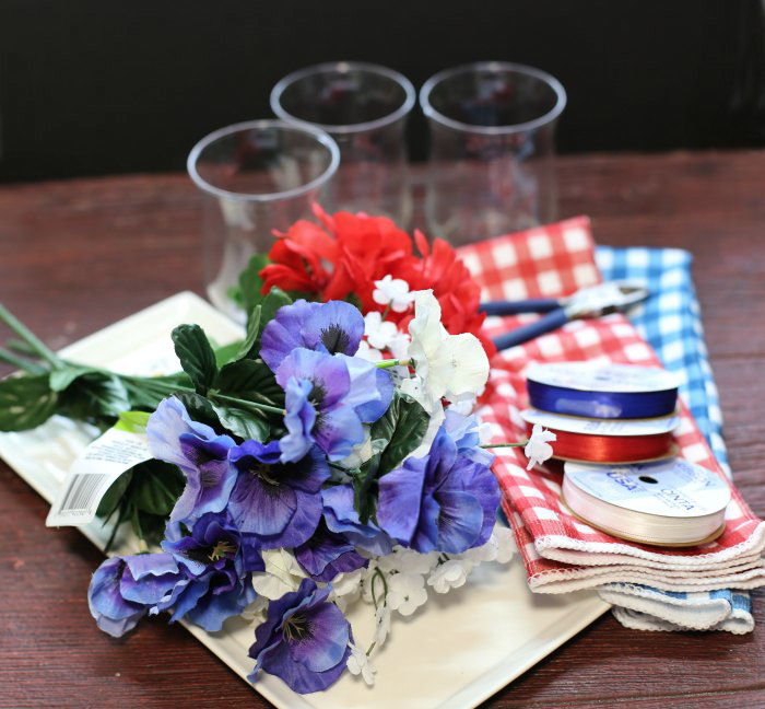 glasses, silk flowers, plate, towels, ribbon and wire snips Supplies for the patriotic table centerpiece including faux flowers, ribbon and glasses.