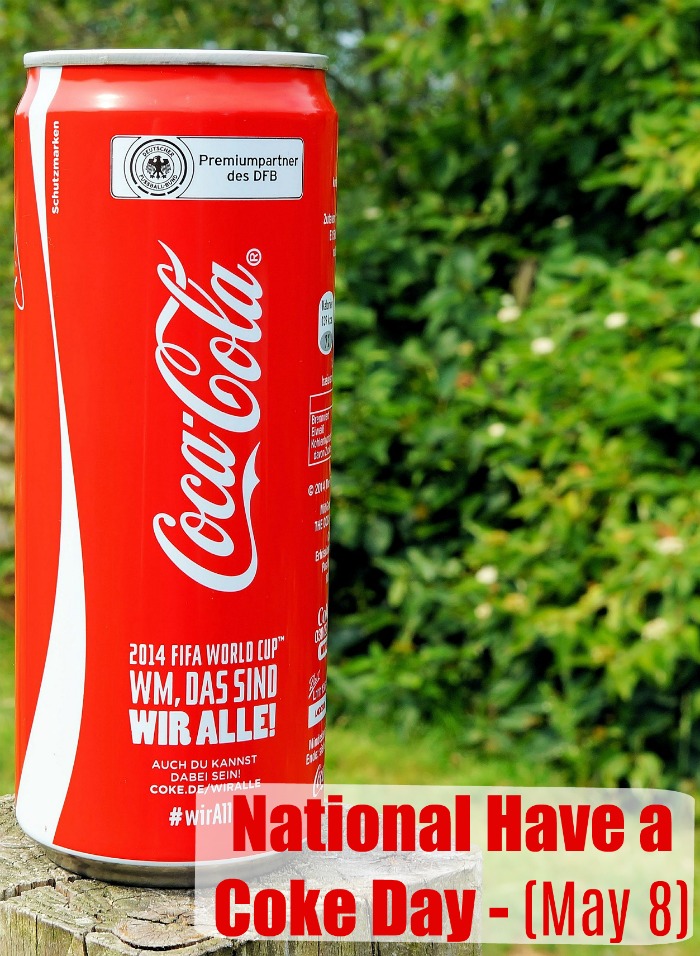 Can of Coke and trees with a text overlay. A tall Coke can on a wooden post in front of a hedge wit a text overlay reading "National Have a Coke Day- May 8".