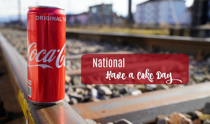 Coke can on a railway with a text overlay. National Have A Coke Day text on a photo of a red can of Coke sitting on train tracks.