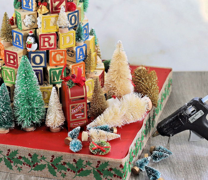 Hot gluing fir trees to a Christmas scene. Faux fir trees, and a glue gun next to an alphabet tree on a wooden base.