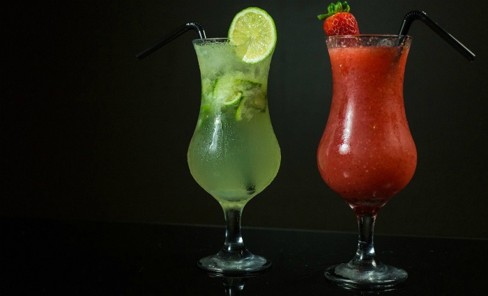 Two cocktails, one red and one green. A green cocktail garnished with a lime and a red cocktail garnished with a strawberry against a black background.