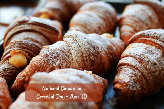 Crescent rolls on a tray. National Cinnamon Crescent Day written in a brown box over a photo of seven cinnamon crescent rolls on a tray.