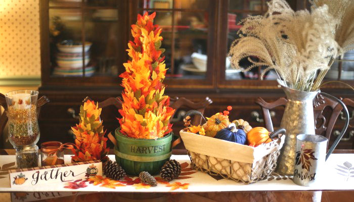 fall vignette Thanksgiving tablescape with basket of pumpkins, silver grass fronds, gather sign, candles, and leaf trees.