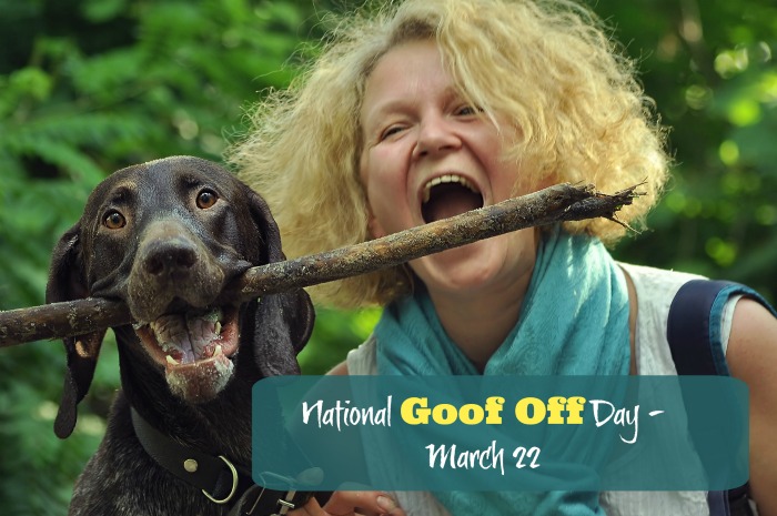 Woman and dog playing with a stick. A dog holding a large stick in its mouth, and a blonde woman joking around and pretending to bite the stick too, with a text overlay reading National Goof Off Day - March 22.