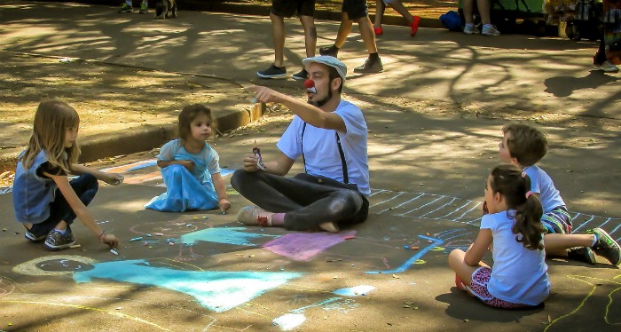 A clown and kids. A man with a clown nose joking around with kids who are drawing with chalk on the pavement.