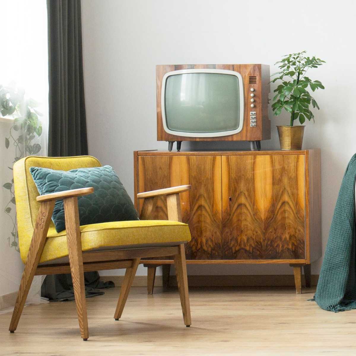 Color TV on a cabinet next to a plant A color TV in a on top of a wooden cabinet with a yellow chair the the left of it under a window with a hanging plant trailing in from the window.