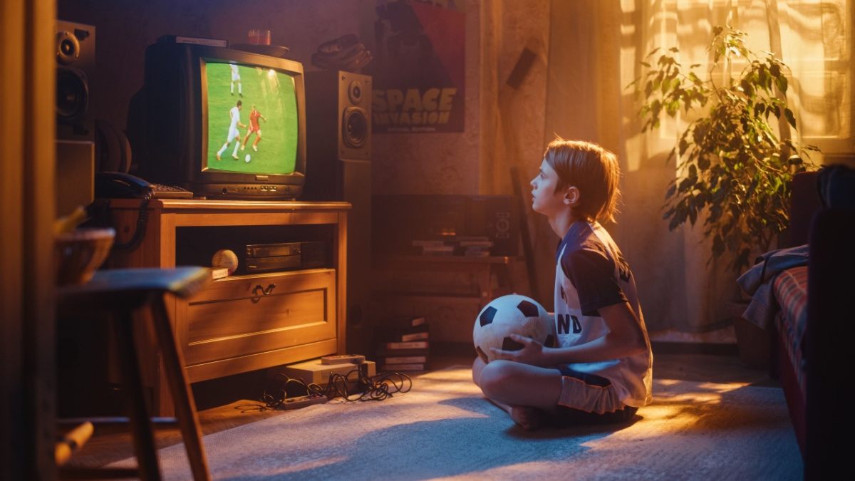 Boy watching a color TV A little boy sitting cross legged on the floor holding a soccer ball watching a soccer match on a retro color tv which is sitting on top of a cabinet in a living room.