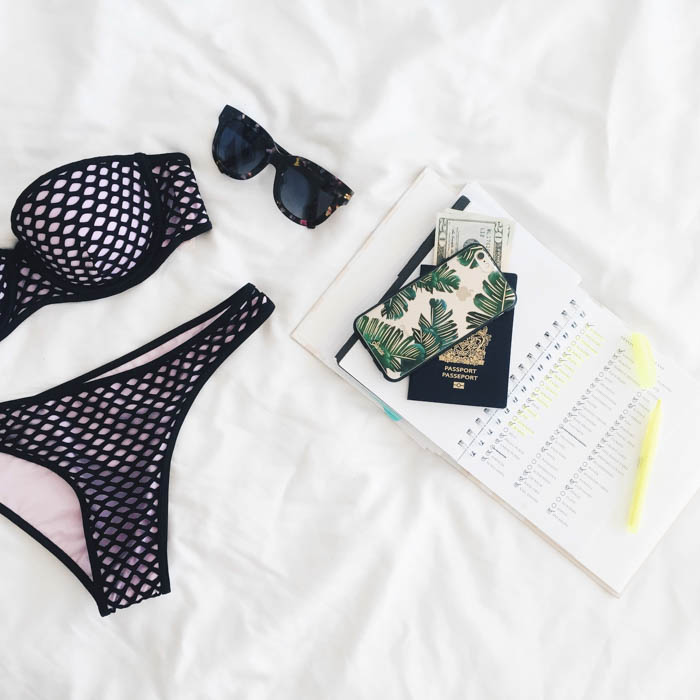 A flatlay photo featuring a black and white mesh bikini, tortoise shell sunglasses, a phone, money, passport, and travel bucket list bikini flatlay, sunglasses, passport