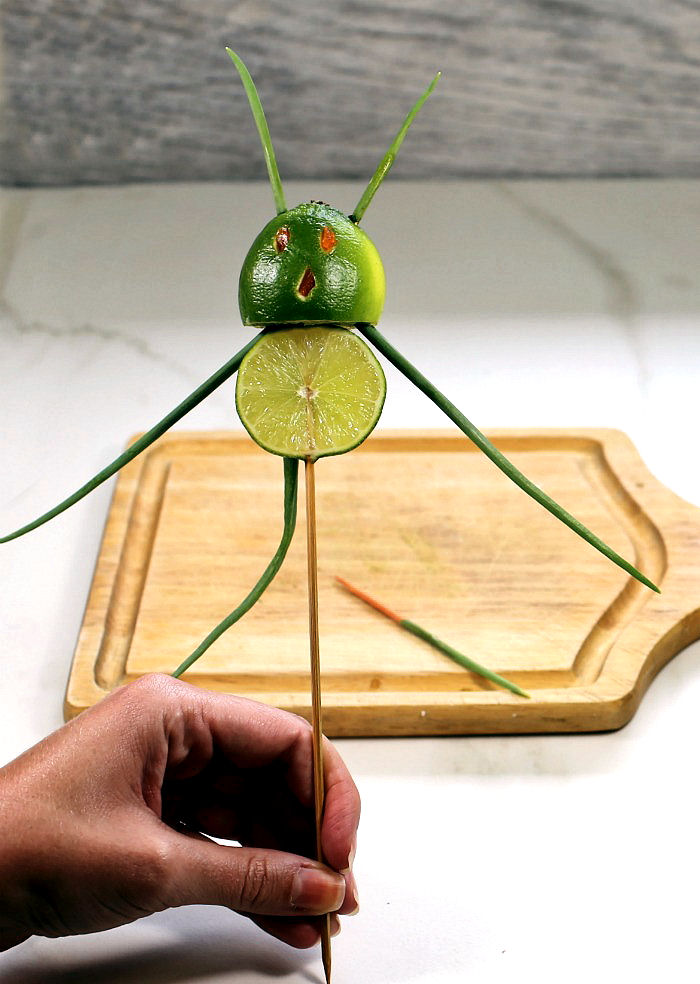 hand holding a lime devil garnish near a cutting board.