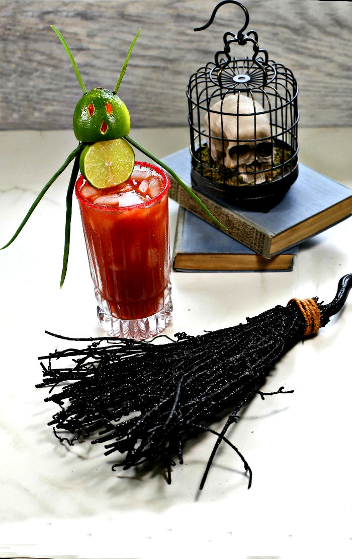Witches broom and skull near a glass of bloody Mary with a lime devil garnish.