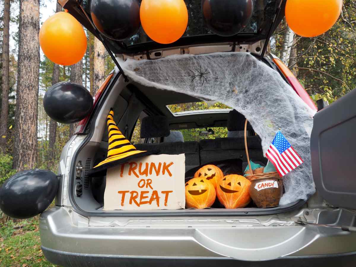Trunk or treat car decorations A grey car with a side opening rear door filled with Halloween car decorations, including a sign that reads "Trunk or Treat" with a black and yellow striped witches hat on it, next to three fake jack o lanterns, and a wicker basket of candy with an American flag sticking out of it, with the outline of the trunk adorned with black and orange balloons and fake spiderweb netting.