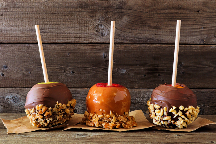 Two candy apples and a caramel apple coated in chocolate and peanut toppings. Two candy apples and a caramel apple coated in chocolate and peanut toppings, sitting on brown paper against a rustic wooden backdrop.