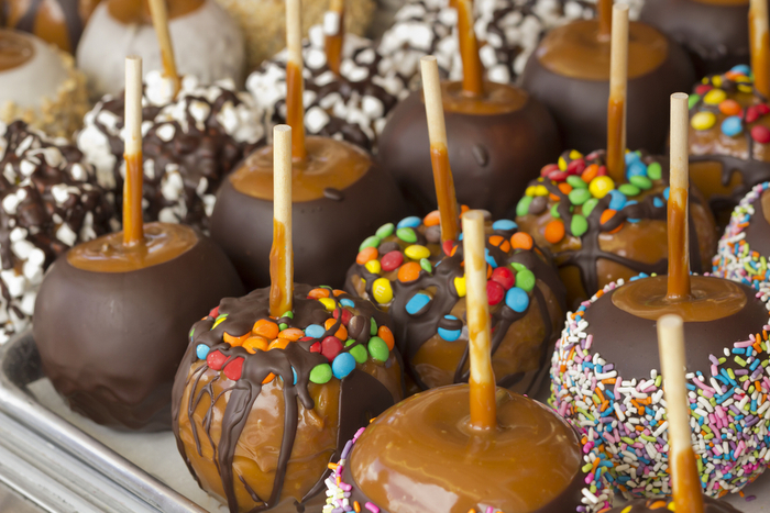 A display of caramel apples with different toppings on each row of caramel apples. An overheard view into a display case of caramel apples with different toppings including M&Ms, sprinkles, nuts, dark chocolate, milk chocolate and white chocolate.