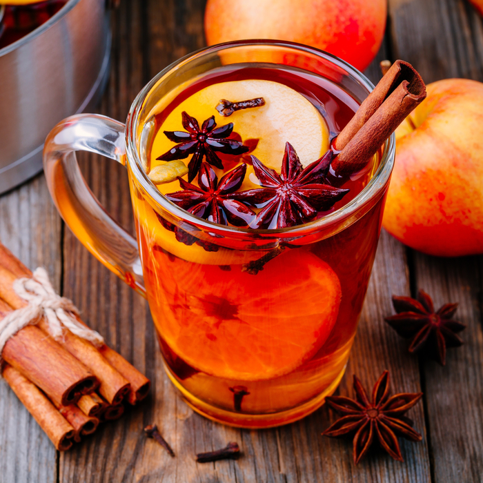 Hot spiced apple cider in a glass mug on a wooden table surrounded by cinnamon sticks, star anise, oranges and apples.