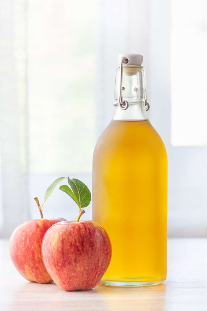 A bottle of apple cider next to two red apples on a table. A bottle of non-alcoholic apple cider on a table with two red apples beside it.