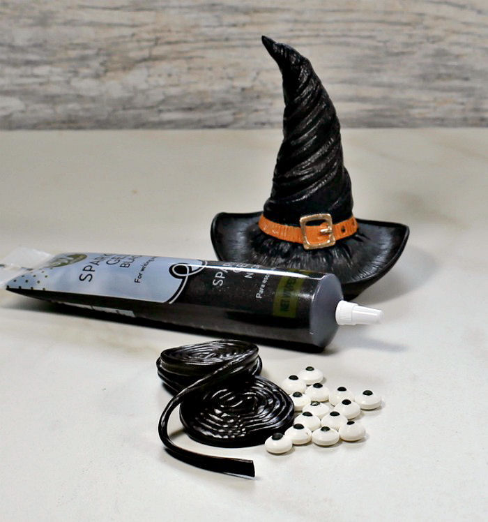 Supplies to make Halloween spider garnishes A witch hat sitting behind the ingredients needed for making a Halloween drink garnish, which include black gel frosting, licorice wheels and edible eyes.