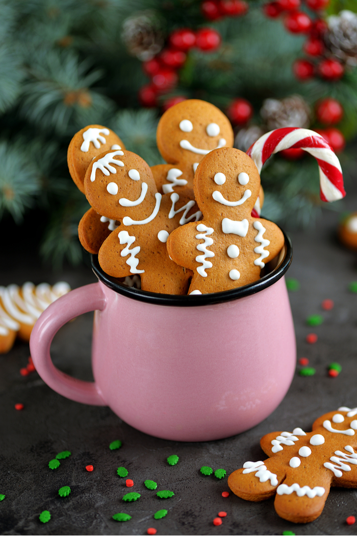 Four gingerbread cookies in a pink mug. Four gingerbread cookies and a candy cane in a pink mug in front of a backdrop of Christmas tree branches.