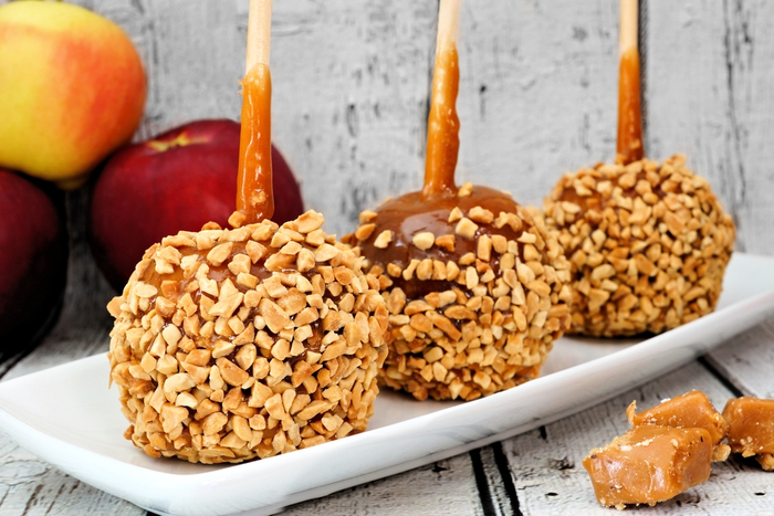 Three peanut coated caramel apples. Three caramel apples covered with peanut toppings on a plate with plain apples and caramels in the background and foreground.