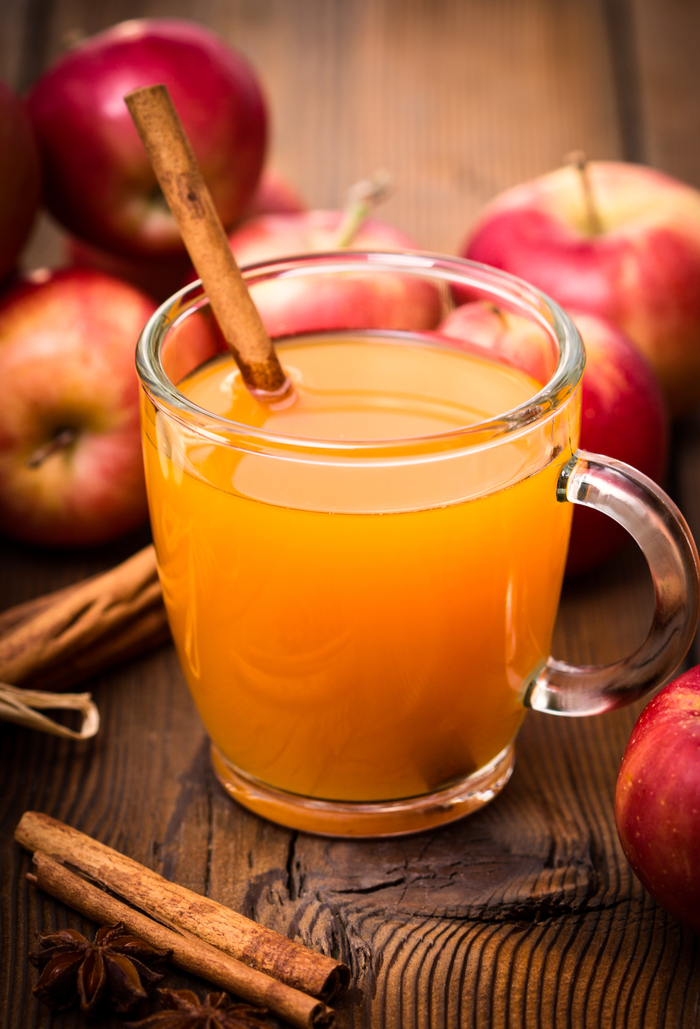 A glass of hot apple cider surrounded by apples and cinnamon sticks. A glass of hot apple cider garnished with a cinnamon stick next to fresh apples on a wooden table.