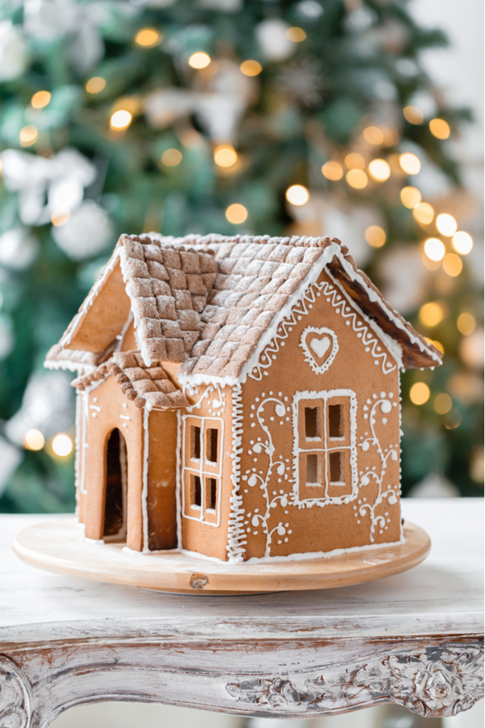 A simply decorated gingerbread house on a wooden table against a Christmas tree backdrop.