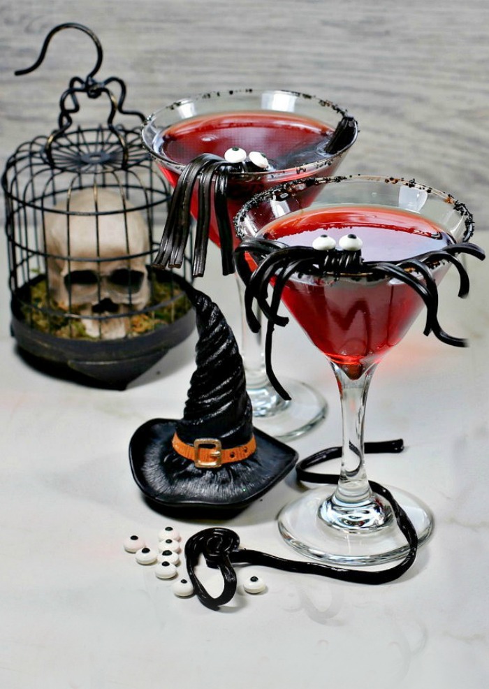 Spider cocktails with drink garnishes Two red Halloween cocktails with spider garnishes, positioned beside pieces of Halloween decor which include a skull in a cage and a witches hat.