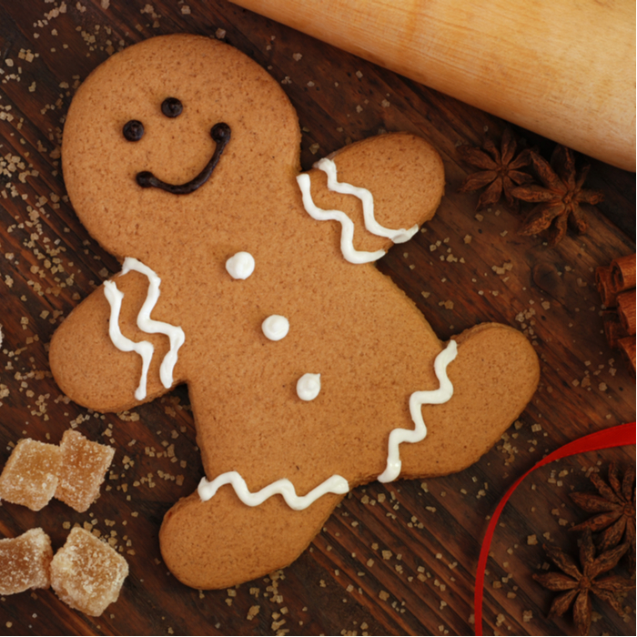 A closeup shot of a gingerbread cookie on a wooden table next to a rolling pin, ginger candies, star anise and a red ribbon.