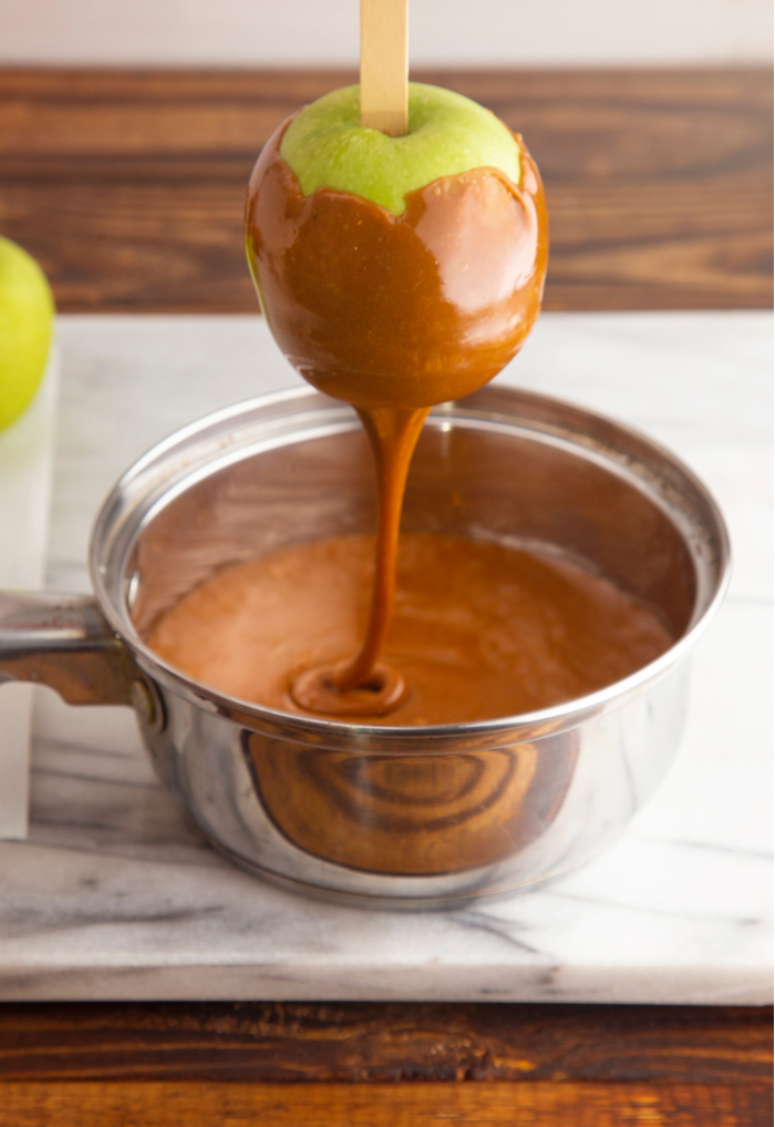 A granny smith apple being dipped into caramel. An apple being dipped into caramel, turning caramel apple ingredients into the finished dessert.
