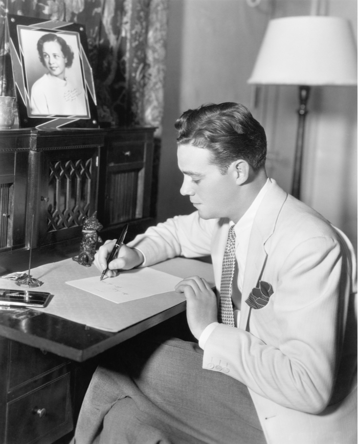 An old black and white photo of a man wearing a suit, sitting at a desk writing a letter.