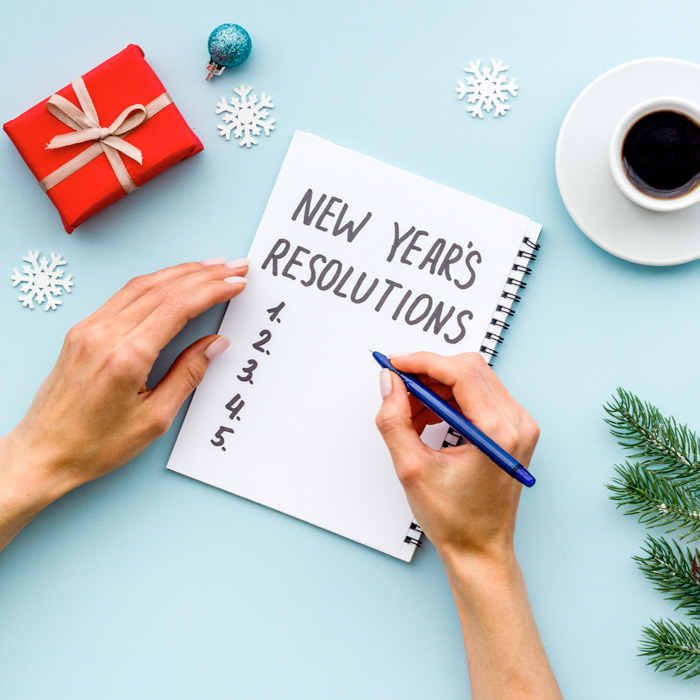 A person writing their New Year's Resolutions with various items in the background, a present, coffee and a Christmas tree branch.