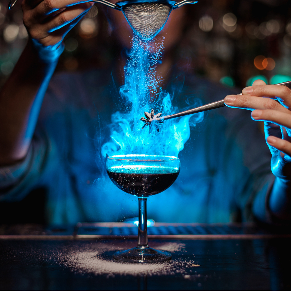 In honor of World Bartending Day, and image of a bartender making a blue cocktail lit on fire to celebrate fun bartending facts.